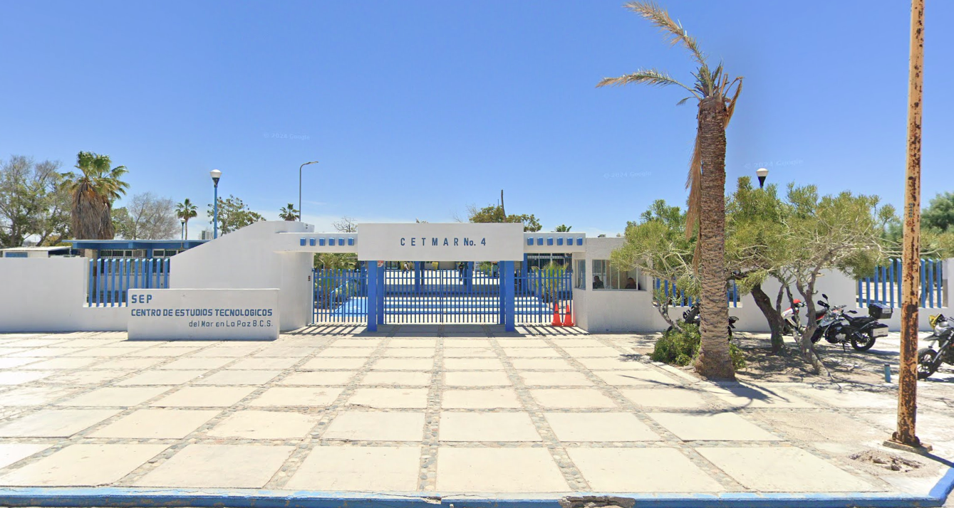 Fotografía en la que se muestra una explanada, al terminar la explanada se encuentra la entrada al plantel, conformada por muros blancos y rejas de color azul y sobre ellas en letras grandes azules dice CETMAR NÚMERO 4. 